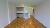 This single-floor unit presents an open-concept layout that combines the living area and kitchen into one cohesive space. The kitchen is neatly tucked into a recessed alcove, fitted with light wood cabinetry, a gas range with a stainless steel finish, a double-basin sink with a dark countertop, and a full-size refrigerator that stands at the boundary between the kitchen and the living area. Under-cabinet space and upper cabinetry provide practical storage. The living area is spacious and bright, featuring classic parquet hardwood flooring laid in a basket-weave pattern that adds warmth and texture underfoot. Neutral white walls keep the interior feeling clean and open. Flush-mount ceiling lighting illuminates both the main living space and a hallway visible in the background, which leads to additional rooms. The hallway suggests further private spaces beyond the main area. Overall, the unit offers a straightforward, functional design with timeless flooring details and a practical kitchen setup suited for everyday living.