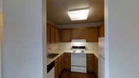 This single-floor space features a compact, efficiently laid-out kitchen that maximizes its available footprint. The kitchen is defined by warm-toned oak cabinetry that lines the walls on multiple sides, offering generous upper and lower storage throughout. The cabinetry pairs with light-colored countertops, creating a classic, understated aesthetic.

Underfoot, rich dark hardwood flooring adds warmth and depth to the space. Overhead, a large flush-mount light fixture provides bright, even illumination across the work areas.

The kitchen is equipped with a full suite of white appliances, including an electric coil range with oven, a dishwasher, and a refrigerator, all neatly integrated into the cabinetry layout. A stainless steel sink is positioned along one wall with an under-counter range hood above the stove providing ventilation. The U-shaped configuration of the kitchen ensures that all key work zones — cooking, cleaning, and storage — are within easy reach of one another, making the space practical and convenient for daily use.