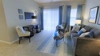 This single-story space centers around a well-appointed living area designed for both comfort and functionality. The room is anchored by large sliding glass doors fitted with vertical blinds that allow an abundance of natural light to fill the space, complemented by an additional side window with decorative blue patterned draping panels that add a refined visual element.

The living area features soft wall-to-wall carpeting in a neutral tone, creating a warm and inviting atmosphere. A coordinated seating arrangement includes a gray sofa with decorative pillows, an accent chair, and a classic round coffee table topped with a distinctive blue gourd vase and floral arrangement, tying the room's color palette together seamlessly.

A dedicated workspace or media area is positioned along one wall, housing a wall-mounted flat-screen television and a dark-toned desk with a matching chair, offering a practical area for work or entertainment. The walls are painted in a muted, calming gray-green tone, enhancing the overall serene ambiance of the space. The combination of natural light, tasteful furnishings, and cohesive décor gives the space a polished and livable character.