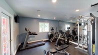 Located at 800 Walden Dr in Wilmington, NC, this single-level fitness center is a well-equipped exercise facility designed to support a comprehensive workout routine. The space is finished with cool-toned, blue-gray walls and durable rubber flooring throughout, creating a clean and functional environment suited for regular use.

The cardio section is stocked with multiple treadmills, an elliptical, an arc trainer, and a recumbent bike, providing a wide range of aerobic training options. For strength training, the facility includes a multi-function cable machine, a squat rack, a dumbbell rack with a full set of weights, and a wall-mounted exercise mat for stretching and floor work.

Full-length mirrors line one wall, visually expanding the space while allowing users to monitor their form during exercise. Recessed lighting illuminates the room evenly from above, and windows allow natural light to filter in, offering views of the surrounding greenery. A wall-mounted flat-screen TV is positioned near the entrance for entertainment during workouts.

The space is accessed through a glass-paned door, and posted fitness center rules ensure the facility remains well-maintained. Security cameras are also present. The overall design is practical and thoughtfully arranged to maximize usable floor space for a variety of fitness activities.