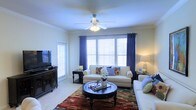 This single-floor residence offers a thoughtfully designed living space that blends comfort and style. The living room serves as the heart of the home, showcasing a bay-style window arrangement dressed with white plantation shutters and rich navy blue curtain panels that frame the natural light beautifully. Crown molding runs along the ceiling, lending an elegant architectural detail to the room, while a ceiling fan with integrated lighting ensures year-round comfort. The space is finished with plush carpeting underfoot, adding warmth and softness to the environment. Furnishings are classic and inviting — a pair of cream-toned sofas anchor the seating area around a dark wood round coffee table, complemented by a vibrant area rug that adds a pop of color and texture. A matching dark wood media console provides a dedicated entertainment area along one wall. Accent lamps and decorative touches, including fresh floral arrangements and throw pillows, contribute to a cozy yet refined ambiance. The layout feels open and airy, with ample room to move and relax. Overall, this residence presents a clean, well-maintained interior with timeless design choices suited for comfortable everyday living.