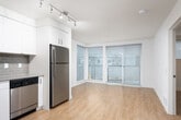 This contemporary single-floor unit offers a clean, modern design throughout, highlighted by warm hardwood-style flooring that flows seamlessly from room to room. The open-concept living and dining area is bathed in natural light, courtesy of expansive floor-to-ceiling corner windows and a sliding glass door that opens onto a private balcony, offering views of the surrounding neighborhood. Track lighting accents the space, complementing the bright, white walls for a crisp, airy atmosphere.

The kitchen is thoughtfully designed with white flat-front cabinetry extending to the ceiling for ample storage, stainless steel appliances including a range, over-the-range microwave, dishwasher, and refrigerator, along with a neutral-toned tile backsplash and a stainless steel sink. The streamlined aesthetic makes it both functional and visually appealing.

The bedroom is a comfortable, well-proportioned room featuring a ceiling fan with integrated lighting, a window with blinds, and the same warm flooring found throughout the unit.

The bathroom is finished with a full soaking tub and shower combo with classic subway tile surround, a vanity with a stone-look countertop and undermount sink, and a large mirror. A stacked washer and dryer unit is conveniently integrated into the bathroom, providing in-unit laundry.