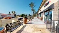 This single-story retail center sits prominently along Foothill Boulevard, offering a well-designed, pedestrian-oriented streetscape in a sun-drenched Southern California setting. The exterior architecture features warm brick cladding accented by stone detailing, arched entryways, and black canvas awnings that lend the center a polished, cohesive aesthetic.

A generously wide concrete promenade runs the length of the storefronts, providing ample space for foot traffic and outdoor activities. The walkway is thoughtfully landscaped with mature palm trees, drought-tolerant ornamental grasses, and striking specimen plants such as aloe trees, all set within mulched planting beds that add texture and visual interest throughout the center.

Large terracotta-style decorative urns serve as focal points along the promenade, complementing the warm tones of the building materials. An outdoor dining patio area features seating shaded by vibrant red umbrellas with decorative palm frond trim, creating an inviting al fresco atmosphere for patrons.

Street-facing retail storefronts are equipped with large glass windows that maximize visibility and natural light. The center benefits from direct boulevard exposure, with convenient surface parking and accessible pathways. The overall ambiance is bright, open, and welcoming, reflecting a contemporary community retail destination designed for comfort and ease of use.