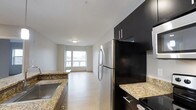 This single-floor residence offers a thoughtfully designed layout that maximizes its comfortable footprint. The kitchen serves as a focal point, outfitted with sleek dark cabinetry that contrasts beautifully against light granite countertops. Stainless steel appliances, including a full-size refrigerator and range, provide a modern, functional workspace ideal for everyday cooking.

The kitchen flows seamlessly into the open living area, creating a connected and versatile space suited for both relaxing and entertaining. Light hardwood-style flooring runs throughout, lending warmth and visual continuity across the unit. Neutral-toned walls keep the interior bright and adaptable to a variety of décor styles.

A window at the far end of the living area allows natural light to filter in, enhancing the overall sense of openness. The clean, contemporary aesthetic is maintained throughout, with modern flush lighting and crisp white ceilings that contribute to an airy atmosphere. The combination of dark kitchen finishes and light living spaces creates a balanced, stylish contrast throughout this well-designed residence.
