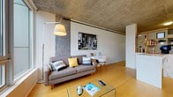 Located at 415 SW Montgomery St in Portland, Oregon, this single-floor urban unit offers a compelling blend of industrial and contemporary design. The exposed concrete ceiling is a defining architectural feature, lending the space a raw, loft-like character that is balanced by warm light hardwood flooring throughout the main living areas.

The living area is anchored by floor-to-ceiling windows that frame sweeping views of the Portland cityscape and surrounding hills, flooding the space with natural light. A partial wall with open built-in shelving divides the living zone from the sleeping area, maintaining an open feel while providing functional definition between spaces.

The kitchen features flat-front bamboo cabinetry paired with clean white countertops, a stainless steel sink, electric cooktop, and oven—delivering a streamlined, modern aesthetic. Under-cabinet lighting adds both practicality and ambiance.

The bathroom is finished in a clean, minimalist style with light tile flooring, a wood-toned vanity, and a full tub-shower combination. The entry area provides a transition point with convenient proximity to the bathroom.

Track lighting runs along the concrete ceiling, complementing the industrial aesthetic while providing flexible illumination throughout the space. Overall, the unit presents a cohesive urban living environment with thoughtful design details.