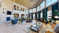 This expansive two-story community clubhouse is designed to offer a comprehensive range of amenities in a polished, well-appointed setting. The centerpiece is a dramatic double-height grand lounge, characterized by soaring ceilings, floor-to-ceiling windows dressed with teal drapes, and an open upper-level balcony that overlooks the main living area below. Comfortable seating arrangements, colorful area rugs, and glass-top coffee tables create an inviting social atmosphere bathed in natural light.

Adjacent to the lounge is a fully equipped kitchen featuring granite countertops, rich wood cabinetry, a black refrigerator, and a bar-height island with seating — ideal for gatherings and events. The fitness center is comprehensively outfitted with treadmills, stationary bikes, cable machines, free weights, kettlebells, stability balls, resistance bands, and floor-to-ceiling mirrors, supported by industrial-style ceiling fans.

A dedicated screening room provides a large projection screen and themed carpeting for a true cinematic experience. A gallery-style hallway showcases framed artwork along its carpeted corridor, lending a curated, refined feel. Private office spaces with tall windows and natural light round out the offerings, providing functional workspace in an elegant environment. The consistent use of grey-blue tones, warm gold ceilings, and white trim details unifies the design throughout.