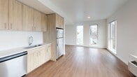 This well-appointed single-floor unit offers a clean, modern aesthetic throughout, characterized by crisp white walls, warm wood-tone accents, and an abundance of natural light. The open-concept living area is anchored by durable wood-style flooring and a generous arrangement of large windows that frame neighborhood views and fill the space with daylight. A wall-mounted HVAC unit ensures year-round comfort.

The kitchen is thoughtfully designed with light wood cabinetry extending to the ceiling for ample storage, white countertops, a textured tile backsplash, and a full suite of stainless steel appliances including an electric range, over-the-range microwave, dishwasher, and refrigerator. The layout flows seamlessly into the living area, making the space feel open and connected.

Two bedrooms are carpeted for comfort and each features sliding closet doors offering generous storage. One bedroom includes a tall window with exterior views, bringing in natural light. Both bathrooms are finished in a clean, minimalist style with wood-style flooring, a full tub-shower combination, and white fixtures. A dedicated in-unit laundry area with stacked washer and dryer adds everyday convenience. The overall design is modern, functional, and move-in ready.