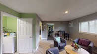 Located at 2294 Zink Rd. in Fairborn, Ohio, this single-floor facility is designed to provide guests with a functional and comfortable communal experience. The space is finished throughout with light tile flooring in common areas and neutral gray and green accent walls that lend a clean, welcoming atmosphere.

The lounge area serves as a central gathering space, furnished with multiple gray upholstered armchairs arranged around a round wooden coffee table. A window with horizontal blinds allows natural light to filter in, and a wall-mounted air conditioning unit ensures year-round comfort. The seating arrangement is relaxed and open, making it ideal for conversation or quiet relaxation.

Adjacent to the lounge, a dedicated fitness room offers a practical workout environment with carpeted flooring and green-painted walls. The room is equipped with a treadmill, elliptical trainers, and a weight bench, along with a wall-mounted flat-screen television. Recessed lighting and a wall-mounted AC unit keep the space comfortable during exercise.

A separate bar-style seating area features a long granite-look countertop lined with cushioned bar stools, illuminated by overhead track lighting. Decorative open wall shelves add a touch of character, and a vending machine provides convenient access to refreshments. Together, these spaces create a well-rounded facility suited for both leisure and fitness.
