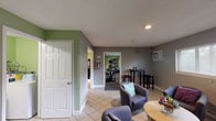 Located at 2294 Zink Rd. in Fairborn, Ohio, this single-floor facility is designed to provide guests with a functional and comfortable communal experience. The space is finished throughout with light tile flooring in common areas and neutral gray and green accent walls that lend a clean, welcoming atmosphere.

The lounge area serves as a central gathering space, furnished with multiple gray upholstered armchairs arranged around a round wooden coffee table. A window with horizontal blinds allows natural light to filter in, and a wall-mounted air conditioning unit ensures year-round comfort. The seating arrangement is relaxed and open, making it ideal for conversation or quiet relaxation.

Adjacent to the lounge, a dedicated fitness room offers a practical workout environment with carpeted flooring and green-painted walls. The room is equipped with a treadmill, elliptical trainers, and a weight bench, along with a wall-mounted flat-screen television. Recessed lighting and a wall-mounted AC unit keep the space comfortable during exercise.

A separate bar-style seating area features a long granite-look countertop lined with cushioned bar stools, illuminated by overhead track lighting. Decorative open wall shelves add a touch of character, and a vending machine provides convenient access to refreshments. Together, these spaces create a well-rounded facility suited for both leisure and fitness.