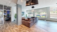 Located at 2521 Kingston Pike in Knoxville, Tennessee, this single-level community lounge offers a generously proportioned, open-concept layout designed for recreation and socializing. The space is finished with wood-look luxury vinyl plank flooring throughout, complemented by a neutral color palette of soft grays and whites that creates a clean, modern aesthetic.

The living and lounge area is anchored by a large, plush sectional sofa positioned beneath a ceiling-mounted projector and large flat-screen TV — ideal for group viewing. An additional wall-mounted flat-screen TV provides entertainment from multiple vantage points. Potted indoor plants add a touch of greenery and warmth to the space.

The game room section features a full-size pool table with a vibrant orange felt surface and a ping pong table, both bathed in natural light streaming through expansive floor-to-ceiling windows and glass doors that lead to an outdoor patio area. High-top bar tables with seating provide a casual gathering spot adjacent to the gaming area.

A dedicated café-style seating zone with round bistro tables and modern chairs offers a quieter space for conversation or work. Large roller shades on the windows allow for flexible light control throughout the day. The drop ceiling with recessed lighting ensures even illumination across the entire floor, while the open sightlines between zones make the space feel connected and inviting.
