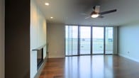 This single-floor space showcases a clean, contemporary design defined by a seamless blend of functional elegance and modern finishes. The living area is anchored by a sleek, linear gas fireplace set flush into a smooth white wall, offering a minimalist focal point that complements the overall aesthetic. Rich, dark hardwood flooring runs throughout, adding warmth and depth to the otherwise crisp interior palette.

Recessed lighting is thoughtfully integrated into the ceiling, providing even, ambient illumination, while a modern ceiling fan with a built-in light fixture enhances both comfort and style. The space is flooded with natural light through expansive floor-to-ceiling sliding glass doors, which frame unobstructed panoramic views of the surrounding landscape. These large-format glass panels create a strong visual connection between the interior and the outdoors, giving the space an open, airy feel.

The neutral gray walls and clean architectural lines contribute to a refined, understated atmosphere throughout. The sliding doors lead to what appears to be an outdoor terrace or balcony, further extending the livable space and enhancing the sense of openness. Overall, the design prioritizes light, views, and modern simplicity.