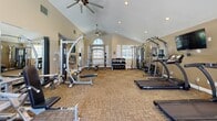 This single-floor fitness center offers a comprehensive workout environment designed to accommodate a wide range of exercise needs. The main workout area is characterized by soaring vaulted ceilings with ceiling fans, recessed lighting, and an arched window that allows natural light to fill the space, creating an open and energizing atmosphere. Warm neutral-toned walls and carpeted flooring throughout contribute to a comfortable and inviting ambiance.

The facility is well-stocked with an extensive selection of equipment, including treadmills, ellipticals, a stair climber, and rowing machines for cardiovascular training. Strength training options include cable machines, multi-station weight systems, a dedicated free weights area with a full dumbbell rack, and kettlebells. A power tower for bodyweight exercises is also available, accompanied by motivational wall signage. A wall-mounted television provides entertainment during workouts.

Connecting to the main area is a dedicated corridor with hardwood-style flooring, housing additional functional training equipment such as medicine balls and stability balls on a storage rack, as well as a water station for hydration. French doors at the end of the corridor offer access to the outdoors, while windows throughout the space provide views of the surrounding greenery and pool area.