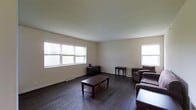This single-floor space presents a clean, straightforward layout centered around a well-lit living area. The room is finished with rich, dark hardwood flooring that contrasts neatly against the neutral, off-white walls and smooth ceiling, creating a balanced and understated aesthetic. Three windows — two larger ones along the left wall and one on the right — allow generous amounts of natural light to fill the room, contributing to an open and airy atmosphere. Baseboard heating runs along the lower edges of the walls, providing a practical and unobtrusive heating solution. The living area is furnished with a dark wood coffee table featuring a lower shelf, complemented by a matching side table and dark upholstered seating, lending the space a cohesive, traditional feel. A small media console sits against the back wall, adding functional storage. The overall ambiance is calm and neutral, offering a versatile foundation that can accommodate a variety of furnishing styles and uses.