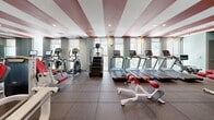 Located at 1023 N Tyndall Ave in Tucson, Arizona, this fitness center offers a comprehensive and thoughtfully designed exercise facility on a single floor. The space makes an immediate visual impression with its striking ceiling design — bold red stripes contrast against white, running the length of the room and lending the space a dynamic, energetic aesthetic that complements its purpose.

The exercise room is generously sized, providing ample room for a wide variety of workout equipment without feeling crowded. Along one wall, a row of large windows brings in abundant natural light and offers views of the surrounding greenery, creating an inviting atmosphere for cardio workouts. This section houses multiple treadmills and elliptical machines, all facing the windows to maximize the outdoor view.

The central and adjoining areas are outfitted with a robust selection of strength-training equipment, including cable machines, resistance stations, and free weight benches finished in a consistent red-and-white color scheme that ties the space together visually. A medicine ball rack and additional functional fitness equipment round out the offerings.

The flooring consists of durable rubber gym tiles throughout, providing safety and comfort underfoot. A water fountain is conveniently mounted along the wall, and glass double doors connect the space to an adjacent common area. Recessed lighting supplements the natural light, ensuring the entire facility is well-illuminated at all times.