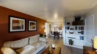 This single-story, 2-bedroom, 1-bathroom residence offers a thoughtfully designed layout that maximizes comfort and functionality. The open-concept living and dining area serves as the heart of the home, creating a seamless flow ideal for both everyday living and entertaining. The living room features warm hardwood-style flooring, a textured ceiling, and a rich terracotta accent wall adorned with framed artwork, adding depth and character to the space. A comfortable seating area anchors the room, complemented by a built-in white shelving unit that provides both display and storage space. The dining area transitions naturally from the living room and is illuminated by an elegant pendant chandelier, creating an inviting atmosphere for meals. The warm tones and cohesive design elements throughout give the space a welcoming ambiance. The two bedrooms offer private retreats, while the shared bathroom provides practical convenience. Consistent hardwood-style flooring runs throughout the main living areas, tying the space together with a clean and modern aesthetic. Overall, this residence balances style and practicality in a compact, efficiently organized single-floor plan.