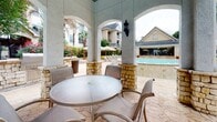 This outdoor amenity area centers around a well-appointed covered pavilion that serves as the primary gathering space alongside a resort-style swimming pool. The pavilion is defined by substantial stucco-finished columns with decorative natural limestone bases, supporting a series of arched openings that frame views of the pool and surrounding grounds. The stamped concrete flooring extends throughout the space, providing a cohesive and durable surface underfoot.

A built-in outdoor kitchen station, constructed with matching limestone masonry, houses a stainless steel grill and provides ample counter space for outdoor cooking and entertaining. Beneath the covered structure, a round glass-top dining table surrounded by patio chairs offers a comfortable seating area with direct sightlines to the pool.

The swimming pool itself is generously sized, with clear blue water and a surrounding deck finished in complementary light-toned pavers. Lounge chairs line the pool deck, and lush landscaping—including palm trees, mature shade trees, and colorful flowering plants—frames the perimeter, creating a vibrant and inviting atmosphere. Iron fencing encloses the entire area, adding both security and a refined aesthetic to this well-designed outdoor retreat.