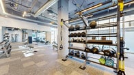 This modern fitness center offers a comprehensive workout environment across a single, open floor. The space features an industrial-chic aesthetic, with exposed ductwork and conduit running along the high ceilings, complemented by linear LED lighting that illuminates the facility throughout. Large floor-to-ceiling windows line the perimeter walls, flooding the interior with natural light and providing views to the surrounding urban environment.

The facility is organized into distinct training zones. A dedicated cardio area houses a row of treadmills and elliptical machines, alongside a wall-mounted flat-screen TV and a multi-station cable machine. Adjacent to this is a free weights zone, equipped with a full rack of dumbbells, adjustable benches, and ample open floor space for functional training. A TRX suspension system is also available for bodyweight exercises.

A well-stocked equipment rack near the entrance features medicine balls, kettlebells of varying weights, and additional fitness accessories. Full-length mirrors along one wall enhance the sense of space and allow users to monitor their form. The flooring throughout consists of durable, patterned carpet tiles designed to withstand high-traffic use. Floor-to-ceiling glass partitions provide a visual connection to adjacent common areas, contributing to the bright, open ambiance of the facility.