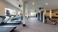 Located on West Rd in Houston, TX, this modern fitness center is a single-floor facility designed to support a comprehensive workout experience. The space is organized into distinct functional zones, each thoughtfully appointed with professional-grade equipment.

The cardio area is generously outfitted with multiple treadmills featuring individual screen monitors, elliptical machines, and stationary bikes, all positioned alongside large picture windows that provide natural light and views of lush outdoor greenery and palm trees. Ceiling fans throughout the space ensure consistent airflow and comfort during workouts.

A dedicated strength and functional training zone features a power rack with weight plates, suspension training systems, wall-mounted pull-up rigs, and spin bikes — making it suitable for a wide range of training styles. Motivational artwork and informational signage, including an "Elevate" fitness program board, add a purposeful atmosphere to this area.

The facility also includes a bank of dark lockers for secure storage, a wall-mounted digital access panel at the entrance, and a built-in audio system. Flooring throughout consists of durable, multi-toned carpet tiles well-suited for gym use. Recessed lighting combined with ample window placement keeps the entire space bright and inviting. The overall design is clean, modern, and functional.