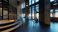 This two-story fitness facility showcases a compelling industrial-chic design, blending raw architectural elements with modern amenities. The lower level commands attention with its soaring double-height ceilings, expansive floor-to-ceiling steel-framed windows, and exposed brick walls that flood the space with natural light and outdoor views. Dark rubber flooring grounds the main workout area, which is equipped with strength machines, free weights, a dedicated training zone with organized equipment racks, a punching bag, and TRX mat stations. A sculptural spiral staircase with wood-and-steel detailing connects the two floors. An entry area features a water station, beverage refrigerator, and curated artwork against the brick backdrop. The upper level contrasts with warm hardwood flooring and an exposed concrete ceiling with visible ductwork and piping, reinforcing the industrial character. Black-framed glass partition walls define dedicated spaces housing Peloton spin bikes, treadmills, and a row of elliptical machines. The upper floor also overlooks the main level through open railings, maintaining visual connectivity throughout. Together, both floors deliver a comprehensive, well-appointed fitness environment with a distinctly urban atmosphere.