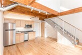 Located at 1703 Cresthill Ave in Royal Oak, Michigan, this two-floor loft-style space offers a striking blend of industrial character and modern finishes. The main level impresses with soaring ceilings clad in warm pine tongue-and-groove planking, supported by rich, dark wood beams and complemented by exposed ductwork that adds an industrial edge. Light hardwood flooring runs throughout, reflecting natural light that streams in through the windows. The kitchen is well-appointed with light maple cabinetry, granite countertops, a white subway tile backsplash, and stainless steel appliances including a refrigerator, dishwasher, and over-the-range microwave. An in-unit stacked washer and dryer provides added convenience. The bathroom features a granite-topped vanity with warm wood cabinetry, a mirrored medicine cabinet, tile flooring, and a porthole-style window that brings in natural light. A staircase leads to the upper mezzanine level, where light pine hardwood floors, clean white walls, and an open railing overlooking the main floor create a bright, airy loft atmosphere. This upper level also features a private entry door. The overall ambiance is modern yet warm, combining natural wood tones with contemporary finishes throughout.