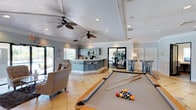 This single-floor multipurpose amenity space is thoughtfully designed to accommodate a variety of recreational and social activities. The layout is open and inviting, anchored by a full-size billiards table positioned at the heart of the room, making it an immediate focal point for entertainment. The space features light-toned tile flooring that extends throughout, contributing to a bright and airy atmosphere enhanced by recessed lighting and dual ceiling fans that provide both style and comfort.

A built-in kitchenette and bar area occupies one side of the room, complete with bar-height seating and cabinetry, offering a convenient space for refreshments and casual gatherings. Adjacent to the kitchenette, a dining area with a table and chairs provides a more structured setting for meals or group activities.

Toward the rear of the space, a dedicated fitness zone is visible through a glass-paneled door, housing exercise equipment for residents seeking a convenient workout option. Crown molding and tray ceiling detailing add a refined architectural touch, elevating the overall design aesthetic. Large windows and glass doors along the perimeter allow natural light to flow in while offering views of the surrounding outdoor greenery, seamlessly connecting the interior to the outside environment.