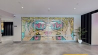 Located at 1514 Hancock St in Quincy, MA, this amenity floor at Nova Quincy is thoughtfully designed to blend bold aesthetics with practical functionality. The welcoming lobby greets visitors with tufted leather seating, mustard yellow accent sofas, polished concrete floors, and glass-partitioned walls that allow natural light to flow freely. A striking pop-art mural featuring bold, colorful graphics serves as a standout visual centerpiece, setting an energetic tone throughout. The "Nova Quincy" neon sign glows above a floral accent wall framed by dramatic black-and-white striped wallpaper, creating an eye-catching entryway. A sophisticated lounge area features curated artwork, teal-framed full-length mirrors, and a mix of plush seating arranged around a sleek conference table — ideal for both relaxation and collaboration. A separate media lounge offers velvet seating in teal and gray tones beneath a large wall-mounted television, surrounded by decorative wallpaper. The fully equipped kitchen includes dark cabinetry, stainless steel appliances, a teal glass backsplash, and ample counter space. The fitness center is well-appointed with treadmills, ellipticals, a rowing machine, a spin bike, free weights, resistance equipment, and wall mirrors — all set against vibrant teal accent walls and durable rubberized flooring.