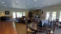 This inviting single-floor space is thoughtfully designed to balance comfort and functionality in an open, flowing layout. The room is anchored by a dramatic floor-to-ceiling stone fireplace, which serves as a stunning focal point and adds a warm, rustic character to the interior. Crown molding along the ceiling line adds a refined architectural detail, while an array of recessed lighting ensures the space is well-lit throughout.

Natural light pours in through multiple large windows and sliding glass doors positioned along the walls, offering views of the surrounding greenery and creating a bright, airy ambiance. The generous proportions of the space allow for distinct functional zones, including a comfortable lounge area furnished with dark leather sofas and a coffee table arrangement, as well as a dining area featuring a classic round dark-wood table surrounded by upholstered chairs.

Warm hardwood flooring transitions seamlessly to patterned area rugs, adding texture and visual interest underfoot. The neutral wall tones complement the rich, dark wood furnishings throughout. A tall wooden armoire provides additional storage and contributes to the space's cohesive, traditional aesthetic. Overall, the design creates a welcoming and versatile environment suited for both everyday use and group gatherings.