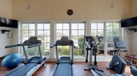 Located at 5412 Edwards Farms Rd in Columbus, Ohio, this single-floor fitness facility offers a bright and functional space designed for community wellness. The standout feature of the gym is its expansive bank of large, multi-pane windows that line the walls, flooding the interior with abundant natural light and providing open views of the landscaped grounds and neighboring community beyond. The warm hardwood flooring adds a polished, inviting aesthetic to the workout environment. The cardio area is well-equipped with multiple treadmills and an elliptical machine, all positioned to take full advantage of the scenic outdoor views — an ideal setup for an engaging and motivating workout experience. The walls feature a neutral, light-toned finish that contributes to the clean and airy feel of the space. A wall clock is mounted centrally, adding a practical touch for those monitoring their workout sessions. The overall ambiance is bright, welcoming, and purpose-built for fitness, combining a functional layout with thoughtful design elements that make the space both comfortable and energizing for regular use.