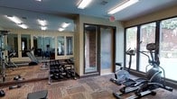 This single-floor fitness room offers a functional and comfortable workout environment designed to accommodate a range of exercise needs. The space features warm beige walls complemented by exposed brick accents surrounding the entryway, giving the room a welcoming, grounded aesthetic. Patterned carpet flooring adds texture underfoot throughout the space.

A full wall of floor-to-ceiling mirrors lines one side of the room, visually expanding the space and providing exercisers with proper form feedback during workouts. A well-organized free weight rack holds a comprehensive set of dumbbells, catering to strength training at various levels. Cardio equipment, including a stationary bike, is positioned near large windows that allow natural light to filter in and offer views of the outdoors, creating an airy and open atmosphere during workouts.

Overhead fluorescent lighting ensures the room remains bright and evenly lit throughout the day. A clearly marked exit door with glass panels provides both safety and additional natural light. The overall ambiance is practical yet comfortable, balancing functionality with an inviting environment suited for regular fitness use.