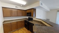 This single-floor residence offers a well-designed layout within a comfortable and functional footprint, featuring one bedroom and one bathroom. The kitchen is a standout space, thoughtfully arranged with warm honey-toned wood cabinetry that extends to the upper walls, providing generous storage throughout. Dark granite-style countertops contrast beautifully with the cabinetry, lending the kitchen a classic, grounded aesthetic. Black appliances, including a gas range with oven and an overhead microwave, are seamlessly integrated into the design. A double-basin sink sits along the extended counter, which wraps to form a convenient breakfast bar or pass-through area, enhancing both functionality and flow. Light hardwood-style flooring runs through the kitchen, contributing warmth and continuity to the space. Recessed ventilation and overhead lighting ensure the kitchen remains bright and well-aired. The open sightlines between the kitchen and the adjacent living area create a connected, spacious feel throughout the residence. Overall, the space combines practical design with a warm, inviting ambiance suited for comfortable everyday living.