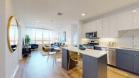 Located at 1 Midtown Bridge St in Hackensack, NJ, this contemporary residence offers a thoughtfully designed open-concept floor plan with high-end finishes throughout. The kitchen is a standout centerpiece, featuring two-tone cabinetry with sleek white upper cabinets and steel-blue lower cabinets, a quartz waterfall island, marble-style backsplash, and a full suite of stainless steel appliances. Light hardwood flooring flows seamlessly into the open living and dining areas, where large windows flood the space with natural light and frame scenic outdoor views of the surrounding landscape. The living room is generously proportioned, offering a relaxed yet sophisticated atmosphere ideal for both entertaining and everyday living. The primary bedroom is a serene retreat, highlighted by a teal accent wall, large windows with sweeping views, and warm wood flooring. A second bedroom features soft sage-toned walls and ample space for a workspace setup. The bathroom is finished with clean white subway tile surrounding the tub and shower, large-format floor tiles, a wood-tone vanity, and a sleek LED-backlit mirror. Recessed lighting throughout adds a polished, modern touch to every room in this well-appointed Hackensack residence.