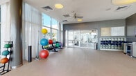 This contemporary fitness center is designed to offer a comprehensive workout experience within a bright, open, and well-appointed environment. The facility is characterized by soaring floor-to-ceiling windows that flood the space with natural light and provide expansive outdoor views, creating an energizing atmosphere for exercise. Ceiling fans and recessed circular light fixtures add to the polished aesthetic throughout.

The cardio zone is generously equipped with Matrix treadmills, elliptical trainers, stationary bikes, and stair climbers, all positioned along the windowed perimeter to maximize natural light and views. A dedicated functional training area features a multi-station power rack complete with a heavy bag, alongside an assortment of colorful stability balls and medicine balls displayed on organized racks.

The strength training section offers a full dumbbell rack with a wide range of weights, an adjustable workout bench, and additional cable and resistance machines. A separate enclosed room, accessible via glass-panel doors, provides additional equipment and workout space.

The support area includes built-in storage cubbies, lockers, a water fountain, and a wall-mounted television, ensuring convenience and comfort for users. The herringbone-patterned carpet and accent walls in a muted gray tone lend a clean, modern finish to the overall design of this well-rounded fitness facility.