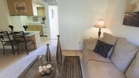 This single-floor residence offers a practical and welcoming layout designed for comfortable everyday living. The living area serves as the heart of the home, featuring neutral-toned carpeting and light-colored walls that create a calm, inviting atmosphere. A glass-topped coffee table anchors the space alongside a plush sofa accented with decorative pillows, while elegant tall vases and a warm table lamp add refined touches to the room's ambiance. A patterned area rug defines the seating zone and introduces subtle texture to the space. The living area flows seamlessly into the kitchen, which is visible through an open passageway. The kitchen features wooden cabinetry in a classic warm tone, a white gas range, and light countertops, offering a functional and well-organized cooking space. The open connection between the kitchen and living area enhances the sense of space and encourages a natural flow throughout the home. A doorway visible from the living area suggests additional rooms beyond, contributing to the overall functional layout of this single-story residence. The consistent use of neutral tones and natural materials throughout ties the space together, resulting in a cohesive and understated aesthetic.