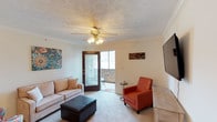 This single-story space centers around a comfortable living area that combines practical design with welcoming details. The room features wall-to-wall carpeting underfoot and is finished with crown molding along the ceiling perimeter, adding a refined architectural touch. A decorative textured ceiling with a centrally positioned ceiling fan and light fixture provides both ambient lighting and airflow throughout the space.

The living area is furnished with a neutral-toned sofa, a black leather ottoman serving as a coffee table, and a vibrant orange accent chair, creating a balanced and functional seating arrangement. A wall-mounted flat-screen television is positioned to the right, complementing the room's layout. Colorful wall art adds a lively touch to the otherwise neutral palette.

A standout feature is the glass-paneled door that opens directly to an outdoor patio, allowing natural light to filter into the room and offering a seamless connection to the exterior. The patio, partially visible through the door, appears to provide a modest outdoor retreat. The overall ambiance is warm and inviting, well-suited for everyday living and relaxed gatherings.