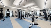 This modern fitness center is a single-floor facility designed to accommodate a wide variety of workout styles and training goals. The space features a clean, contemporary aesthetic with white walls, a drop-tile ceiling fitted with recessed lighting and ceiling fans, and a distinctive blue and white speckled rubber flooring that adds both visual appeal and functional safety underfoot.

The cardio zone is well-appointed with multiple treadmills, elliptical trainers, and stationary bikes, all positioned near large windows fitted with roller shades that allow for natural light while maintaining a comfortable environment. Full-length mirrors are strategically placed throughout to assist with form and to visually expand the space.

The strength training area offers a comprehensive selection of selectorized cable machines, multi-functional power racks, and a full dumbbell rack ranging across a wide variety of weights. Dedicated benches and weight stations are positioned throughout for both isolation and compound movements.

A functional training zone adds versatility to the facility, featuring a multi-station rig complete with a heavy punching bag, TRX suspension trainers, and a large tractor tire for dynamic training. Wall-mounted flat-screen televisions are positioned for convenient viewing throughout the space. Large windows and glass entry doors contribute to an open, airy ambiance, making this fitness center an inviting and fully capable training environment.