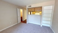 This two-floor space offers a practical and functional layout designed to maximize utility. The main living area is finished with neutral-toned carpet, creating a comfortable and understated foundation for the room. A built-in shelving unit lines one wall, providing ample storage and display space without the need for additional furniture.

A defining feature of the space is the open pass-through kitchen, framed by warm wood-toned upper cabinetry. This design choice creates a visual connection between the kitchen and living area, making the layout feel open and communicative. The kitchen itself is visible from the living space, showcasing additional cabinetry and a sink area beneath a lighted workspace.

An HVAC vent is integrated into the ceiling, ensuring climate control throughout the space. A doorway from the living area leads into a hallway connecting to a bathroom, which is partially visible and features warm lighting and a vanity. The overall color palette is neutral and light, with white walls and trim lending a clean, airy feel. The two-floor configuration provides additional living or storage potential beyond what is visible in the main level.