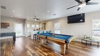 This single-floor recreation and game room is thoughtfully designed to serve as a dynamic entertainment hub. Rich, multi-toned hardwood flooring runs throughout, providing a warm and inviting foundation that ties the entire space together. At the center of the room sits a solid wood billiards table with a blue felt surface, serving as the focal point of the space. Adjacent to it, a foosball table adds an additional layer of entertainment options.

A comfortable lounge area is positioned to one side, anchored by a large sectional sofa in a cool navy hue, accented with teal and earth-tone throw pillows. A wall-mounted flat-screen television and a sleek ceiling fan with integrated lighting complete this relaxation zone. Multiple windows fitted with white blinds allow ample natural light to fill the room while maintaining privacy.

The wet bar area features white cabinetry, a stainless steel sink, and generous counter space, ideal for entertaining. A vibrant, large-scale piece of artwork adds a bold splash of color to the neutral-toned walls. Bar-height seating with teal upholstered chairs complements the bar setup. Recessed lighting and ceiling fans are distributed throughout, ensuring comfort and ambiance across every zone of this versatile, well-equipped space.