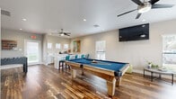 This single-floor recreation and game room is thoughtfully designed to serve as a dynamic entertainment hub. Rich, multi-toned hardwood flooring runs throughout, providing a warm and inviting foundation that ties the entire space together. At the center of the room sits a solid wood billiards table with a blue felt surface, serving as the focal point of the space. Adjacent to it, a foosball table adds an additional layer of entertainment options.

A comfortable lounge area is positioned to one side, anchored by a large sectional sofa in a cool navy hue, accented with teal and earth-tone throw pillows. A wall-mounted flat-screen television and a sleek ceiling fan with integrated lighting complete this relaxation zone. Multiple windows fitted with white blinds allow ample natural light to fill the room while maintaining privacy.

The wet bar area features white cabinetry, a stainless steel sink, and generous counter space, ideal for entertaining. A vibrant, large-scale piece of artwork adds a bold splash of color to the neutral-toned walls. Bar-height seating with teal upholstered chairs complements the bar setup. Recessed lighting and ceiling fans are distributed throughout, ensuring comfort and ambiance across every zone of this versatile, well-equipped space.