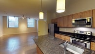 This single-floor space offers a thoughtfully designed open-concept layout that seamlessly connects the kitchen and living areas. The kitchen is a standout feature, showcasing rich dark granite countertops with an undermount sink, warm-toned wood cabinetry, and a full suite of stainless steel appliances, including a French door refrigerator, microwave, and range. A large kitchen island anchors the space, providing ample prep and dining surface. Pendant lighting with cylindrical drum shades hangs above the island, adding both warmth and a modern aesthetic.

The living area extends naturally from the kitchen, benefiting from polished concrete flooring that runs throughout the open space, lending an industrial yet refined character. A ceiling fan with a track lighting system keeps the space well-ventilated and versatile in lighting options. A large window allows natural light to fill the living area, creating a bright and airy atmosphere. Neutral-toned walls complement the warm wood and stone finishes found across the space.

A distinctive accent wall in a soft yellow-green hue adds a touch of color near the transitional hallway, contributing to the overall modern and urban character of this well-appointed single-floor residence.