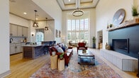 Located at 9451 Welby Rd in Thornton, Colorado, this single-floor space makes an immediate impression with its soaring two-story ceilings and an expansive curved wall of multi-pane windows that floods the interior with natural light and provides sweeping outdoor views. Light hardwood flooring runs throughout, lending warmth and continuity across the open layout.

The main living area is anchored by a sleek, modern fireplace with a rustic wood mantel and a built-in niche beside it that serves as both a firewood storage display and a decorative shelving unit — a standout architectural detail that blends function with character. A coffered tray ceiling detail and an elegant chandelier add refinement overhead, while pendant lighting defines the adjacent wet bar area.

The wet bar is well-appointed with taupe cabinetry, granite countertops, a white subway tile backsplash, a matte black faucet and hardware, and a stainless steel beverage refrigerator — making it ideal for entertaining. A rustic wood-paneled island with a granite top anchors the bar area, complementing the overall warm, Western-inspired aesthetic.

Throughout the space, the design balances contemporary finishes with organic, rustic accents, creating an inviting and cohesive atmosphere. Large windows and an arched glass door further enhance the connection between the interior and the outdoors.