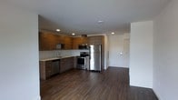 This single-floor space showcases a thoughtfully designed open-concept layout that seamlessly blends the kitchen and living areas into one cohesive environment. The kitchen is a standout feature, outfitted with a full suite of stainless steel appliances including a refrigerator, electric range, built-in microwave, and dishwasher. Warm-toned wood cabinetry lines the walls both above and below the countertops, providing generous storage while lending a contemporary yet inviting aesthetic. A stainless steel sink with a modern faucet is neatly integrated into the countertop along the back wall. Recessed lighting is installed throughout the ceiling, offering clean, even illumination across the space. The flooring is a rich, dark hardwood that runs continuously through the kitchen and into the adjoining living area, adding a sense of warmth and visual depth. The open plan creates a spacious, airy feel, with clean white walls providing a neutral backdrop that complements the wood tones. A private entryway with a solid white door is visible from the main living space, offering a clear sense of separation and privacy. Overall, the space delivers a polished, modern aesthetic suited for contemporary living.