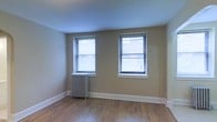 This single-floor space offers a clean, straightforward layout with timeless architectural details. The main room is finished with warm hardwood flooring that runs throughout, adding character and a sense of continuity to the space. Neutral-toned walls provide a versatile backdrop suited to a variety of uses and décor styles.

A standout feature of the room is the classic cast-iron radiator, indicative of traditional building construction and offering reliable radiant heat during colder months. Two double-hung windows dressed with horizontal blinds are positioned along the exterior wall, allowing for generous natural light while maintaining privacy. The white-trimmed windows and baseboards provide a crisp contrast against the warm wall tones, lending the room a polished, well-maintained appearance.

The ceiling is smooth and bright, contributing to an open and airy feel. The overall ambiance is understated yet functional, with the combination of hardwood flooring, period-appropriate heating, and natural light creating a comfortable and practical environment. The space presents a solid foundation for customization, making it well-suited for residential or light professional use.