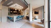 Located at 930 Ridgeview Dr in Allen, TX, this thoughtfully designed single-floor space combines a living lounge and kitchen into a cohesive, upscale environment. The open layout is anchored by large-format light-toned tile flooring that flows seamlessly throughout, creating a clean and expansive feel.

The kitchen area features a substantial bar-height island with a textured concrete finish, topped with smooth quartz countertops and surrounded by teal velvet barstools. Behind the bar, a glossy dark subway tile backsplash, matte black fixtures, stainless steel appliances, and mirrored upper cabinetry add depth and sophistication. Pendant lighting with glass fixtures illuminates the space warmly overhead.

The living area offers multiple seating vignettes, including rich brown leather chairs paired with round white pedestal side tables, and a dark tufted sofa anchoring the central lounge zone. A boldly patterned accent wall with textured wallcovering and a framed wall-mounted TV define one side of the room, while a large-scale automotive artwork lends an artistic focal point. Floor-to-ceiling windows along one wall flood the space with natural light and offer views of the surrounding exterior. The overall ambiance is modern, curated, and inviting — designed for both comfort and visual impact.