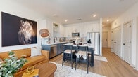 This single-floor residence offers a well-designed open-concept layout that connects its primary living spaces with a clean, contemporary aesthetic. A welcoming entry hallway features light wood flooring, crisp white panel doors with matte black hardware, and a wall-mounted thermostat, setting the tone for the interior's modern design language. A decorative live-edge wood piece adds an organic accent upon entry.

The living area is anchored by large street-facing windows dressed with linen drapes, flooding the space with natural light and offering views of the surrounding cityscape. The room accommodates both a seating arrangement centered around a round wood coffee table and a media wall with a mounted flat-screen television.

The kitchen stands out with its two-tone cabinetry — white upper shaker-style cabinets paired with dark lower cabinets — complemented by a white subway tile backsplash, light quartz countertops, and a full suite of stainless steel appliances. A central island with bar seating provides additional prep space and casual dining.

A dedicated office area features light wood flooring, open metal shelving units flanking a framed abstract artwork, and a dark work desk — creating a focused yet polished workspace. Throughout, the residence maintains a cohesive, modern aesthetic with neutral tones and thoughtful detailing.