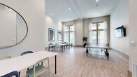 Located in Houston's 77023 neighborhood, this single-floor multi-use space functions as both a dining area and a recreational gathering room. The interior is defined by impressively high ceilings fitted with recessed lighting, which contribute to an open and airy atmosphere throughout. Light-toned hardwood flooring runs the length of the space, complementing the crisp white walls and creating a clean, contemporary aesthetic.

Large, floor-to-ceiling windows dressed with sheer curtains line the perimeter, flooding the room with abundant natural light and offering views of the surrounding streetscape. The dining area features multiple white rectangular tables paired with a mix of navy and sage green chairs, providing flexible seating arrangements suitable for both casual meals and group gatherings. A curated selection of bold, colorful framed art pieces adorns the walls, adding a vibrant visual focal point to the space.

A full-size ping-pong table occupies the recreational zone, complemented by a wall-mounted flat-screen television — making this area equally suited for leisure and entertainment. A potted indoor plant near the windows introduces a touch of greenery. The entryway features glass-paneled doors that reinforce the room's bright and welcoming character. Overall, the space balances functional dining with social and recreational use in a polished, well-lit environment.