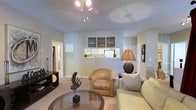 This single-floor residence offers a welcoming living area defined by neutral wall tones and plush wall-to-wall carpeting that contributes to a warm, comfortable atmosphere. The ceiling-mounted fan with integrated lighting provides both air circulation and overhead illumination, while a decorative table lamp adds layered ambient lighting to the space. The living area is furnished with a cushioned sofa and a rounded accent chair, complemented by a circular glass-top coffee table that anchors the seating arrangement. A low-profile media console sits against one wall, providing storage and display space. A decorative sculptural piece adds an artistic touch to the room. A mirrored pass-through connects the living area to the kitchen, offering a glimpse of white appliances and cabinetry beyond. Three framed landscape artworks are displayed above the pass-through, adding visual interest to the wall. Doorways lead to additional areas of the home, including what appears to be a hallway with further rooms. The overall ambiance is calm and cohesive, with a practical layout that balances open living with defined functional zones.