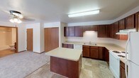 This single-floor unit offers a practical and comfortable layout designed for everyday living. The kitchen is a standout feature, equipped with rich dark wood cabinetry, light countertops, a double-basin sink, a full suite of white appliances including a range, dishwasher, and refrigerator, and a peninsula island that provides additional prep space and a natural divide between the kitchen and adjoining areas. Stone-look tile flooring ties the kitchen together with a clean, durable finish. The open living area is generously proportioned, finished with neutral carpet, and features sliding glass doors that lead to a private balcony overlooking a scenic outdoor setting with views of neighboring rooftops and greenery. A dedicated laundry area is conveniently accessible from the living space. Two bedrooms offer comfortable retreats, each with plush carpet, baseboard heating, and windows that bring in natural light. One bedroom enjoys a particularly expansive window with picturesque views. The unit includes two full bathrooms, both featuring dark wood vanities, light countertops, tub-shower combinations, and stone-look tile flooring. The overall ambiance is clean, neutral, and move-in ready, with warm wood tones providing a cohesive design thread throughout.