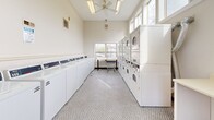Located at 10900 Oak Forest Parkway Dr in Saint Louis, MO, this single-floor communal laundry facility offers a practical and well-organized layout across a comfortable footprint. The space is accessed through a dedicated entrance and hallway, leading into the main laundry area, which serves as the focal point of the facility.

The laundry room is generously equipped with a row of front-loading washing machines along one wall and a bank of stacked dryers along the opposite side, providing ample capacity for multiple users. High ceilings and a ceiling fan contribute to an open, airy atmosphere, while large windows along the perimeter flood the space with natural light and offer views of the surrounding greenery. A folding/sorting table and laundry cart are also present for added convenience. The flooring throughout features a classic grey penny-tile pattern that adds visual interest while remaining durable and easy to maintain.

Two bathrooms are accessible from the main area, each finished with matching penny-tile flooring, warm wood-toned vanity cabinets, mirrored medicine areas with vanity lighting, and white walls. One bathroom includes accessibility grab bars, underscoring the facility's inclusive design. The overall ambiance is clean, bright, and functional.