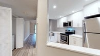 This single-floor unit presents a thoughtfully updated interior with a cohesive, modern design. The open-concept living area is defined by vaulted ceilings that lend a sense of spaciousness, complemented by a ceiling fan with integrated lighting and a track lighting system. Light wood-tone flooring runs continuously throughout, unifying the space with a warm, neutral palette against soft gray walls and crisp white trim.

The kitchen is efficiently designed in an L-shaped layout, featuring white shaker cabinetry, bright white quartz countertops, and a classic subway tile backsplash. A standout stainless steel farmhouse sink anchors the space, accompanied by a full suite of stainless steel appliances including an electric range, over-the-range microwave, and dishwasher. Recessed lighting illuminates the kitchen, which opens to the living area via a raised breakfast bar counter. A window at the far end of the kitchen allows natural light to filter through.

A hallway connects the living areas to the remaining rooms, lined with louvered bifold closet doors that provide storage access while maintaining a clean visual line. A washer and dryer are also tucked conveniently within the hallway. Textured ceilings are present in portions of the unit, adding subtle contrast to the otherwise streamlined interior.