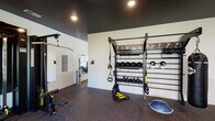 This single-level fitness center is thoughtfully designed to accommodate a wide range of workout styles and fitness goals. The space is divided into distinct training zones, each outfitted with purpose-specific equipment. A dedicated functional training area features a wall-mounted rig stocked with TRX suspension trainers, kettlebells, medicine balls, and resistance tools, providing a comprehensive setup for bodyweight and functional movements. Adjacent to this zone, a pull-up bar and additional suspension equipment extend the training options further.

The strength training section houses a multi-station cable machine, adjustable weight benches, and a full dumbbell rack with a broad weight range, all positioned in front of large wall mirrors that visually expand the space and aid in form monitoring. A wall-mounted flat-screen television adds to the amenity. The cardio zone includes an air bike and a rowing machine, positioned near large windows that offer views of the surrounding outdoor area, allowing natural light to fill the space.

Throughout, dark rubber flooring provides durability and sound absorption, while a dark-painted ceiling with recessed lighting and ceiling fans creates a focused, gym-like atmosphere. The overall layout is open and functional, designed to support both individual training and group exercise.